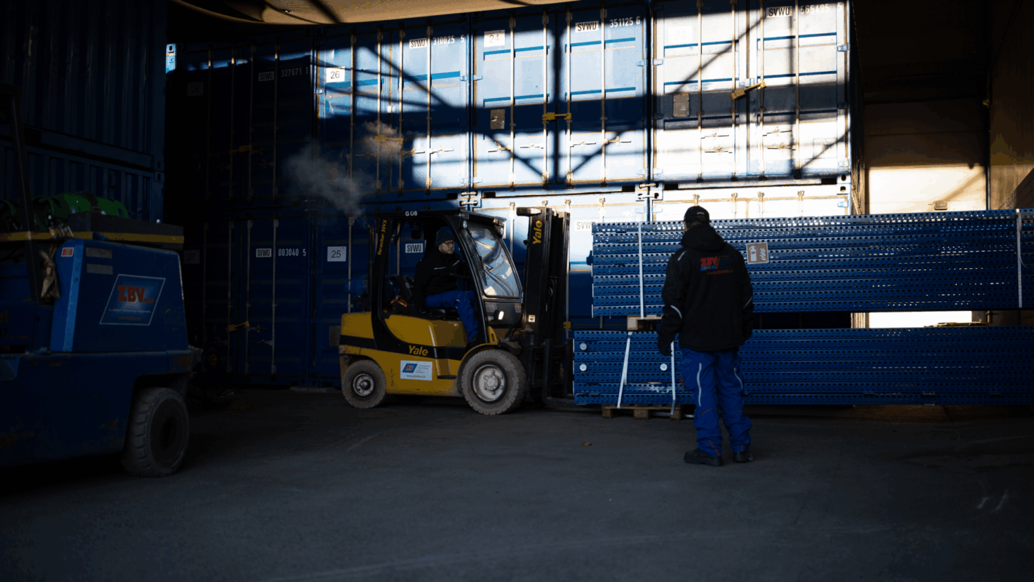 Gabelstaplerfahrer lädt schwere Metallregale vor Containern, während ein weiterer Mitarbeiter die Ladung koordiniert – professioneller Logistik- oder Montageservice in einer Lagerhalle währen Lagerümzuge.