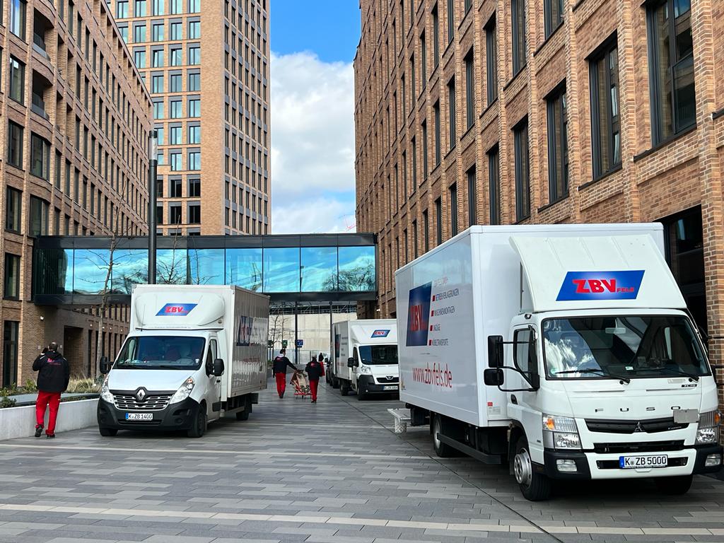 Zwei Lieferwagen in einer urbanen Straße zwischen hohen Backsteingebäuden, Menschen im Hintergrund.