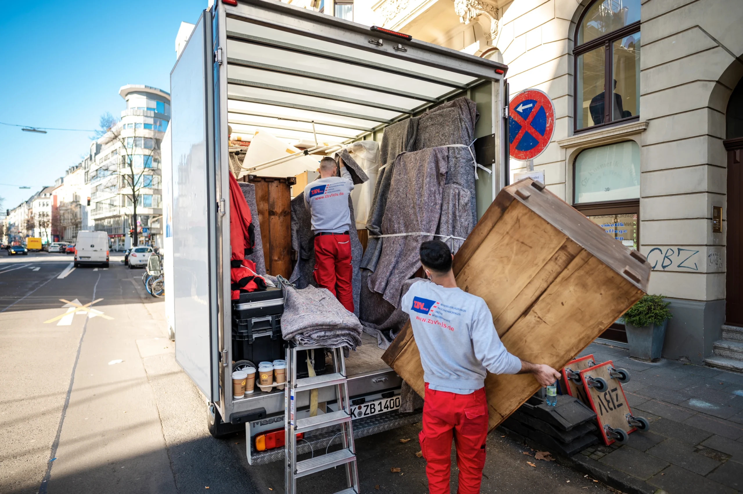 Zwei Umzugshelfer laden Möbel aus einem LKW vor einem Wohnhaus aus.
