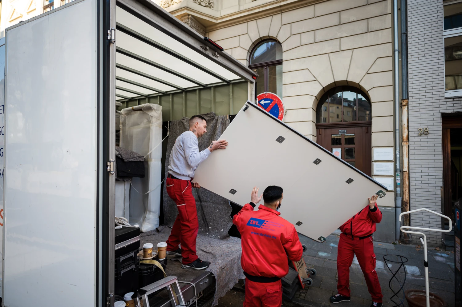 Drei Männer tragen einen großen Kühlschrank aus einem Lieferwagen auf eine Straße vor einem Gebäude.