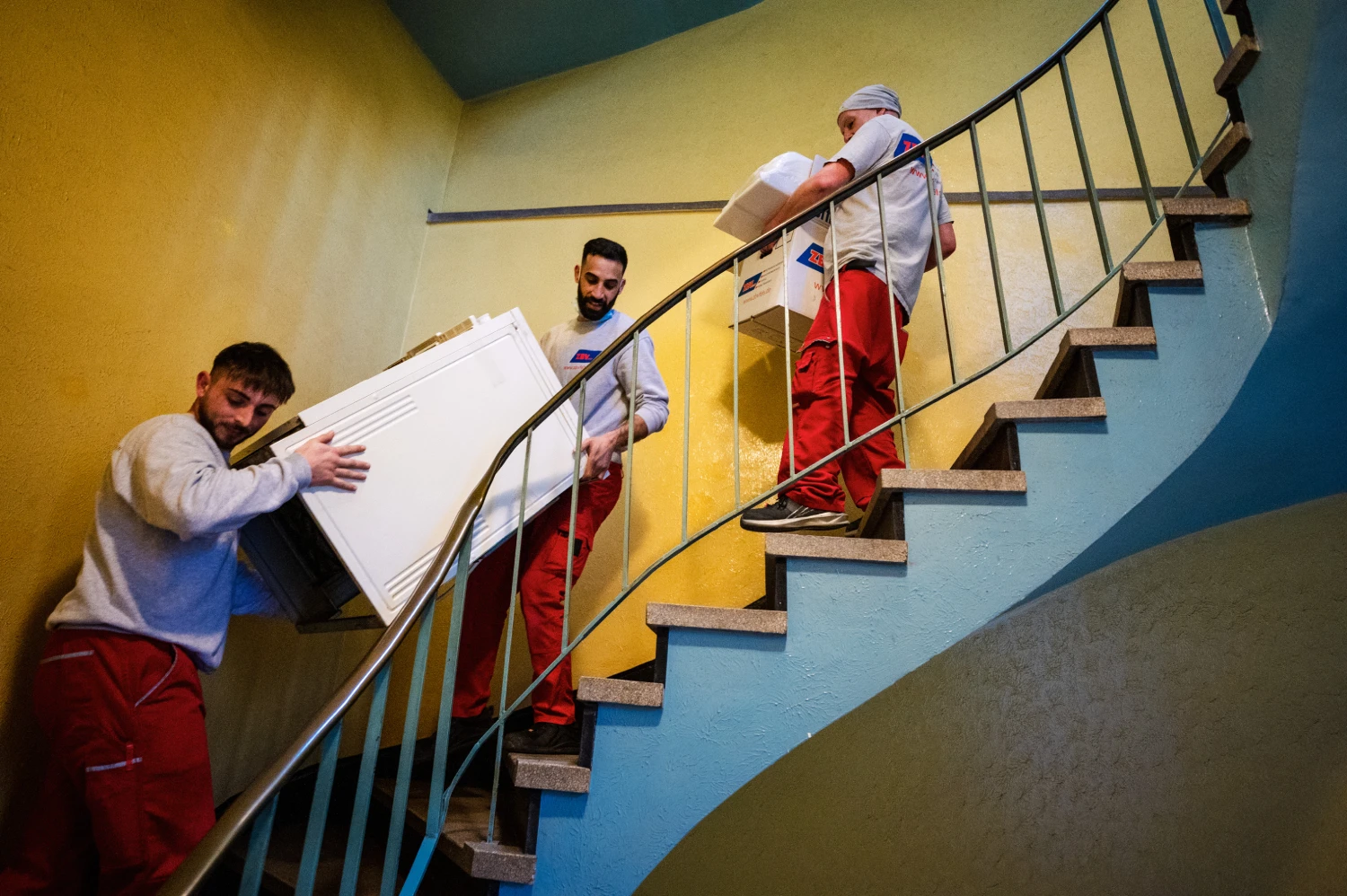 Drei Männer tragen einen großen weißen Gegenstand eine Treppe hinauf.